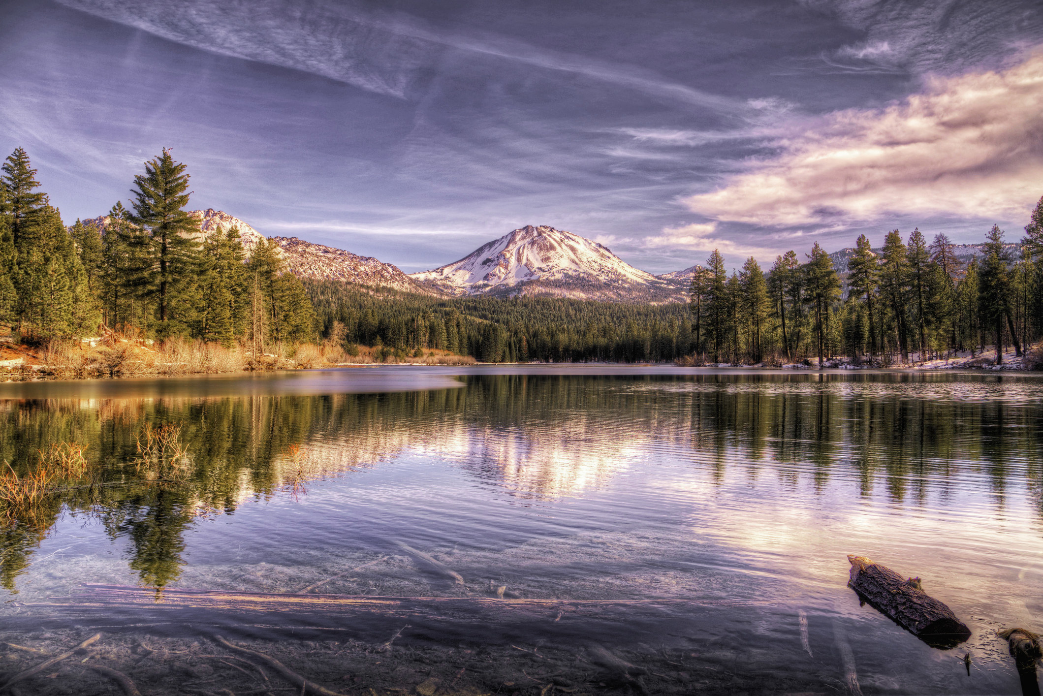 A snowy mountain peak and a glassy lake surrounded by trees in Lassen Volcanic National Park.