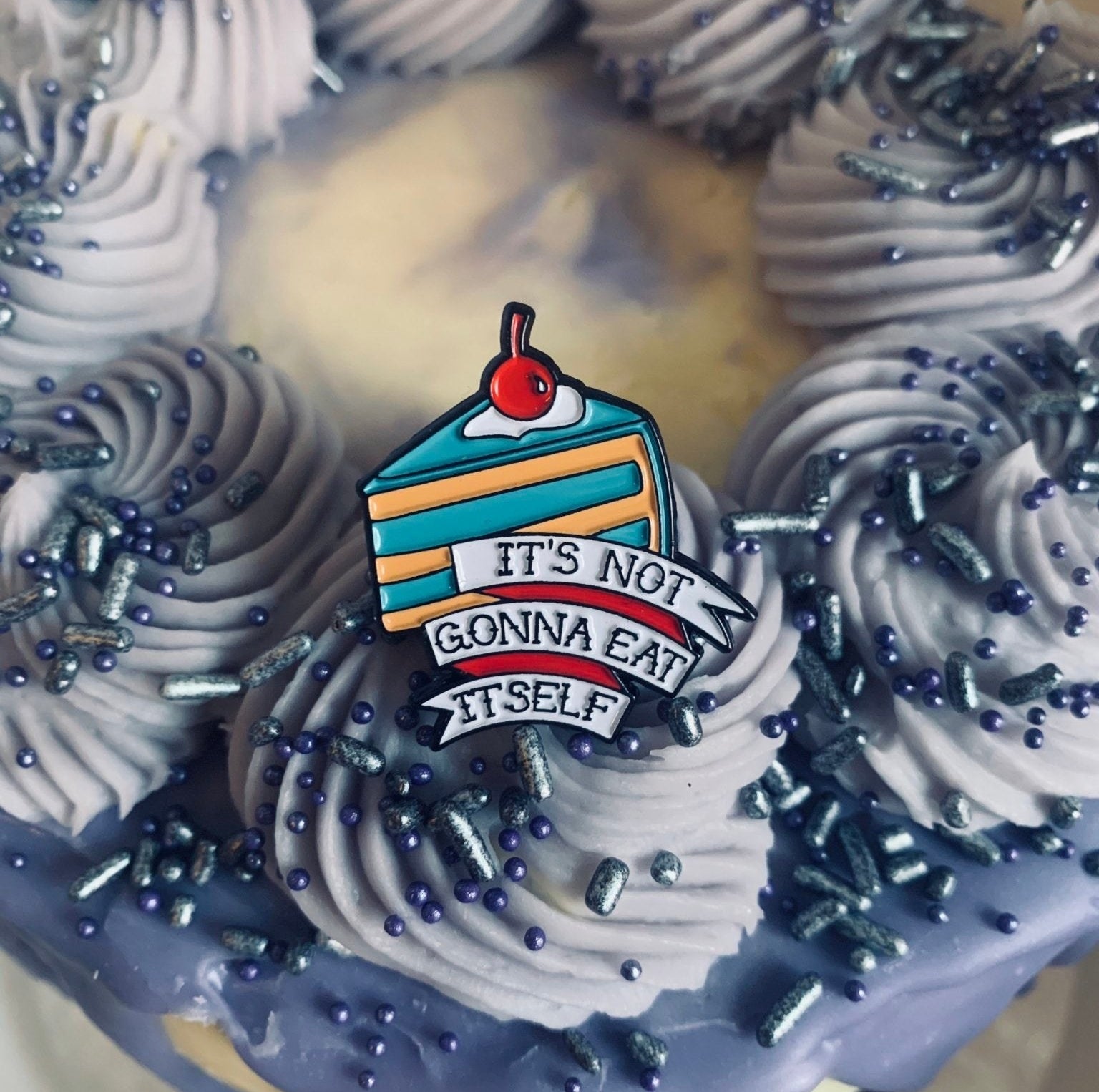 an enamel pin of a blue and yellow slice of cake with a cherry on top that says "it's not gonna eat itself"