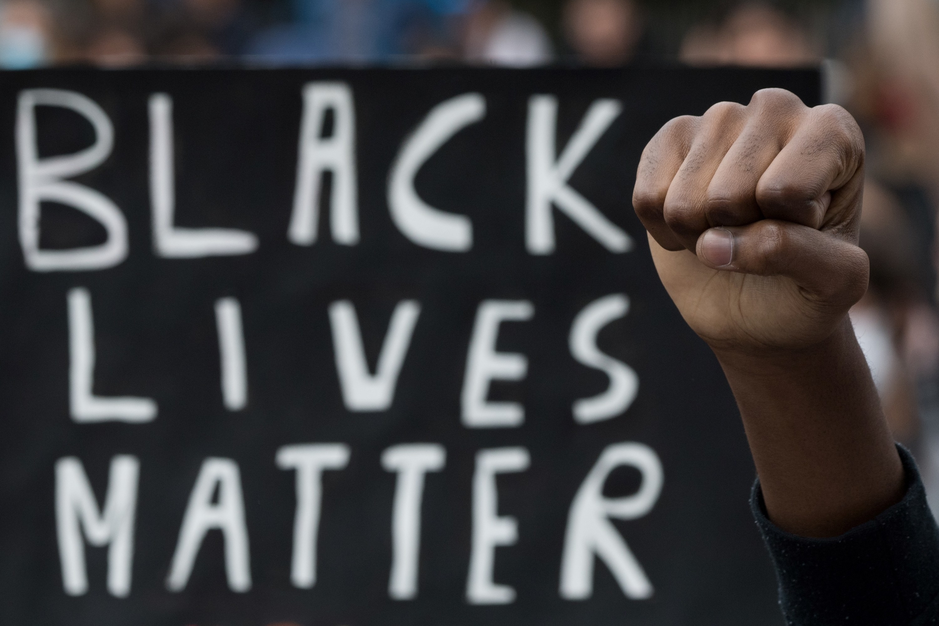 A Black person raises their fist in front of a Black Lives Matter protest sign
黒人の1人が「Black Lives Matter」と書かれた看板の前で、拳を掲げている。