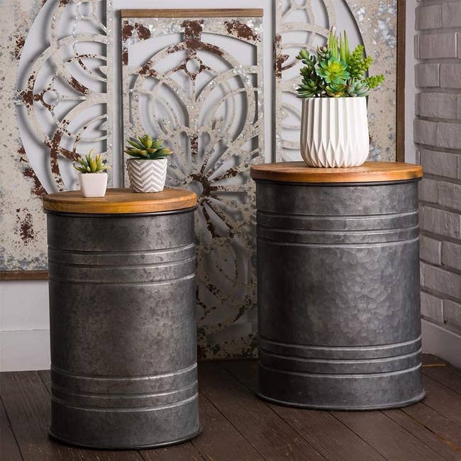 Two metal stools with a vintage milk canister vibe, each artistically worn with dents and discoloration. The lids have a smooth, natural wood finish. 