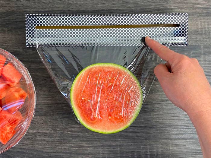 A person using the plastic wrap dispenser to cut cling flip overtop of a halved watermelon.