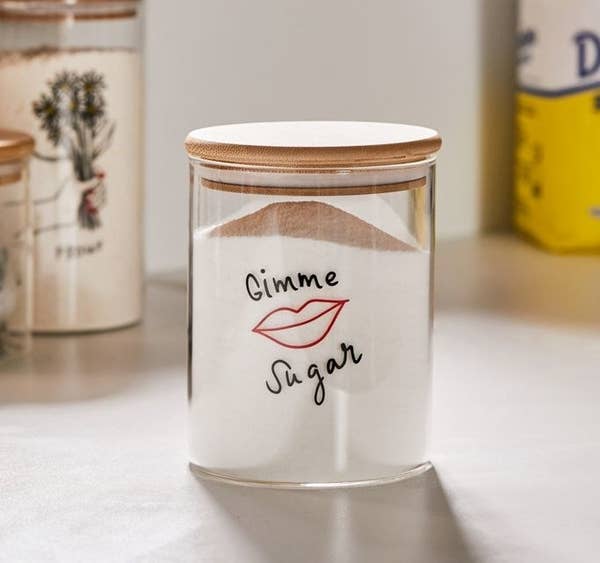 Clear glass jar with a wood lid and an illustration of lips in red and the words "Gimme Sugar" in black above and below it