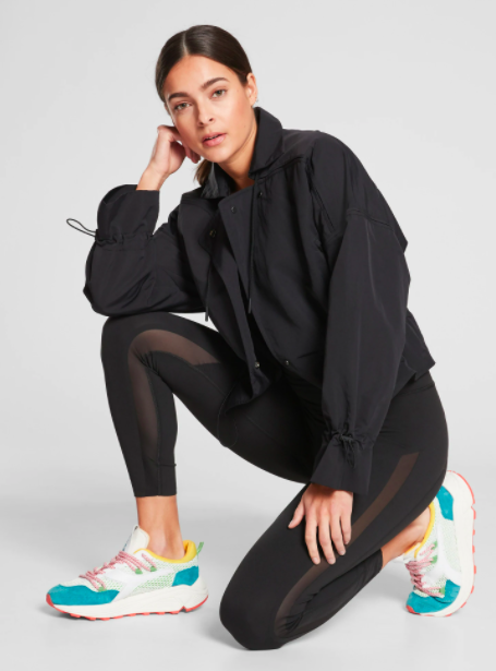 Model wears black Athleta jumpsuit with a black jacket and colorful sneakers