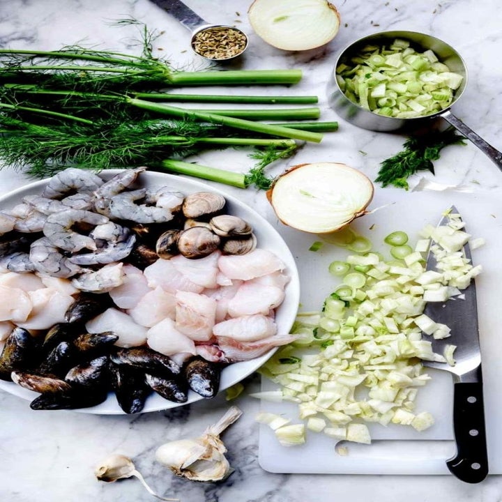 Ingredients for cioppino on a countertop including raw mussels, shrimps, and scallops, garlic, onion, and fennel.