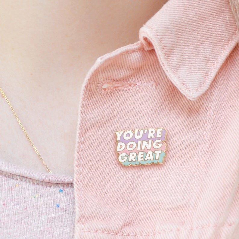 The pin, with white block letters that say &quot;YOU&#x27;RE DOING GREAT&quot; over a pastel purple, pink, and green background, pinned on a jacket
