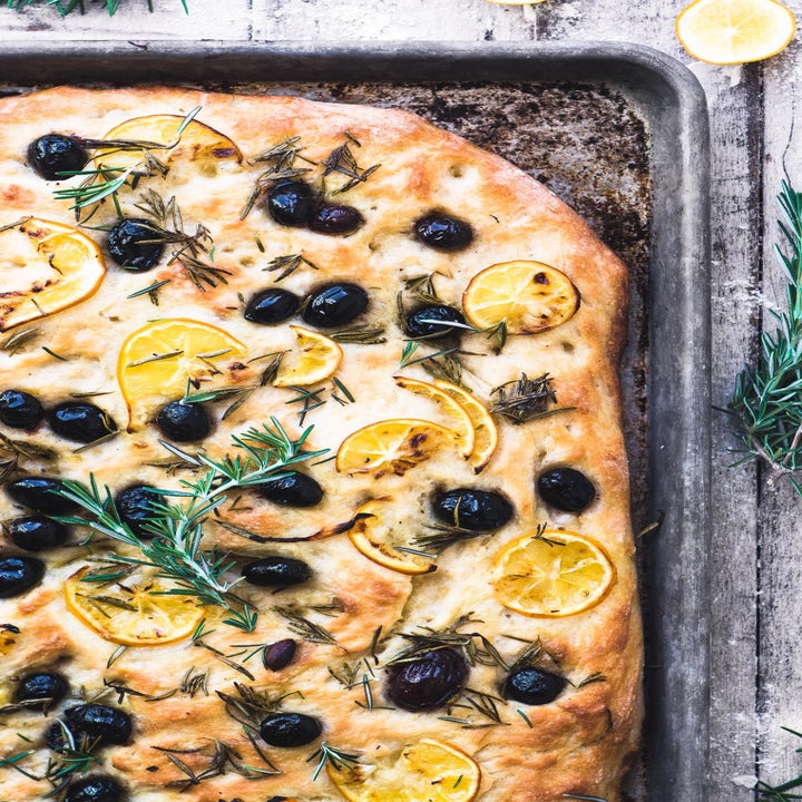 A loaf of baked focaccia bread topped with Meyer lemons, rosemary, and olives.