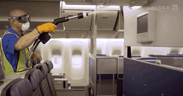 man inside an airplane, wearing protective gear and spraying disinfectant out of a hose onto a TV screen