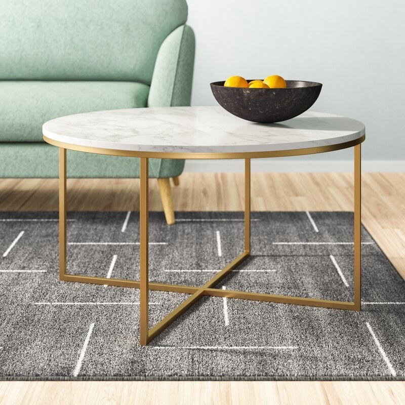 The circular shaped coffee table with a marble-look top and four hollow gold-toned legs that meet in an &quot;x&quot; at the bottom
