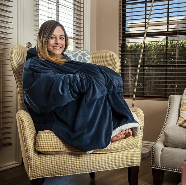 A woman sits curled up on an armchair, wearing The Comfy and smiling at camera