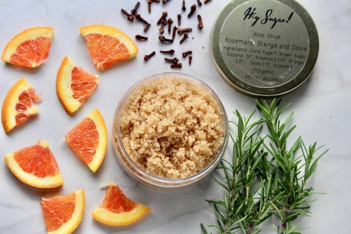 A flat lay shot of the brown scrub in a labeled jar with the lid off and raw ingredients of citrus, rosemary, and clove around it