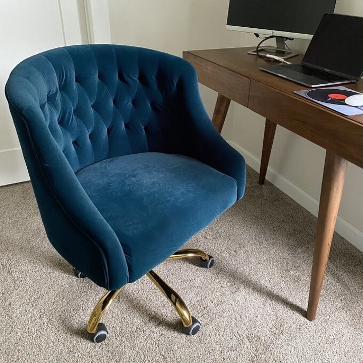 A blue round office chair with a padded velvet and a tufted back. It has golden legs and rolls on wheels