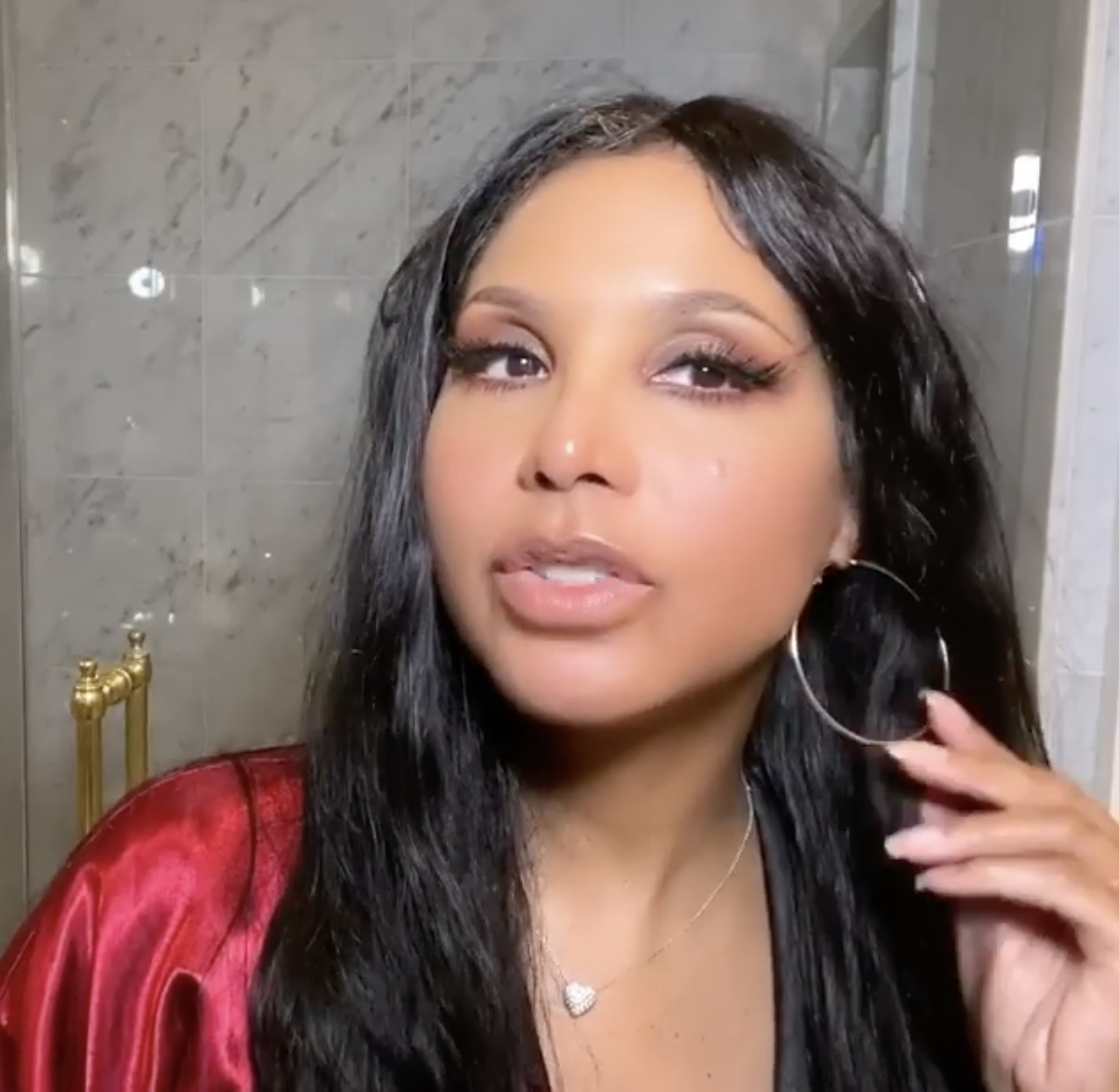Toni Braxton shows off her earrings with makeup on her face in a red robe