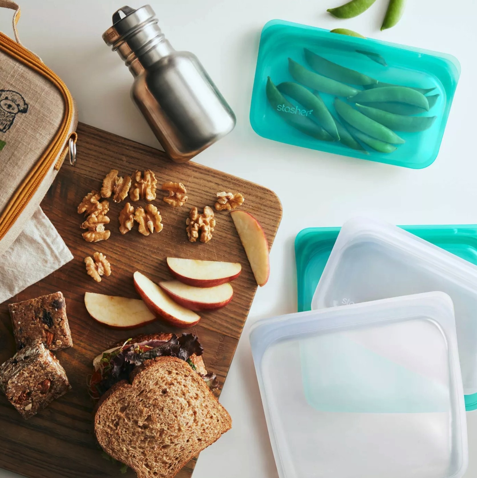 Three empty silicone bags and one silicone bag with edamame next to a chopping board with a sandwich, apple slices, walnuts, and a granola bar.