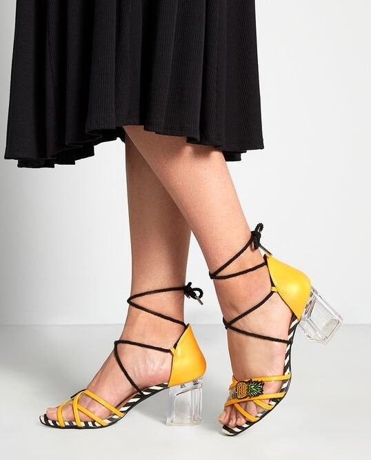 Feet wearing the yellow sandals with a lucite block heel, black and white soles, black tie straps, and pineapple applique details on the clear and yellow toe straps