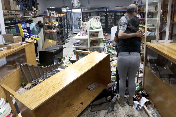 A man hugs his adult daughter amid their vandalized store, where the floors are scattered with bottles and a table is overturned