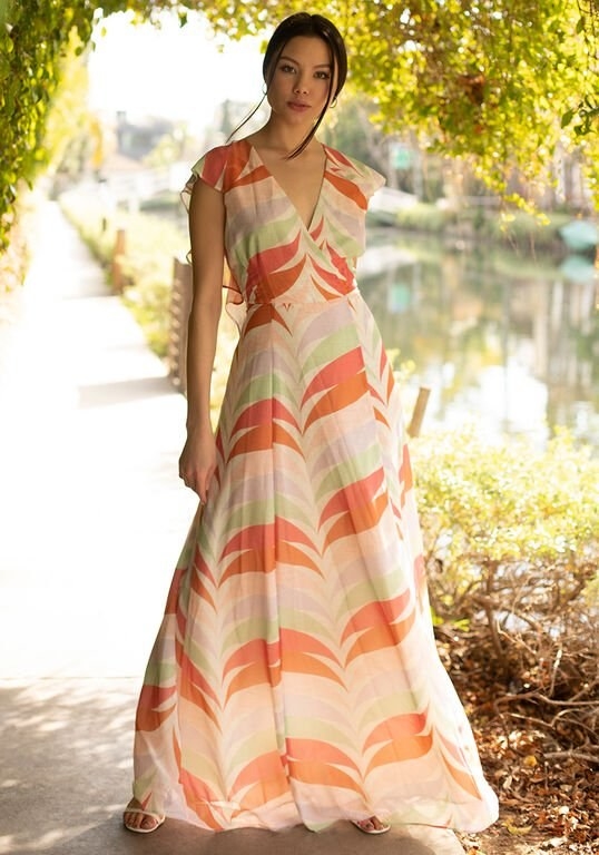 A model in the ruffle-backed, surplice neck, short-sleeve dress that&#x27;s white with an orange, coral, pale green, lilac, and peach feather-shaped print