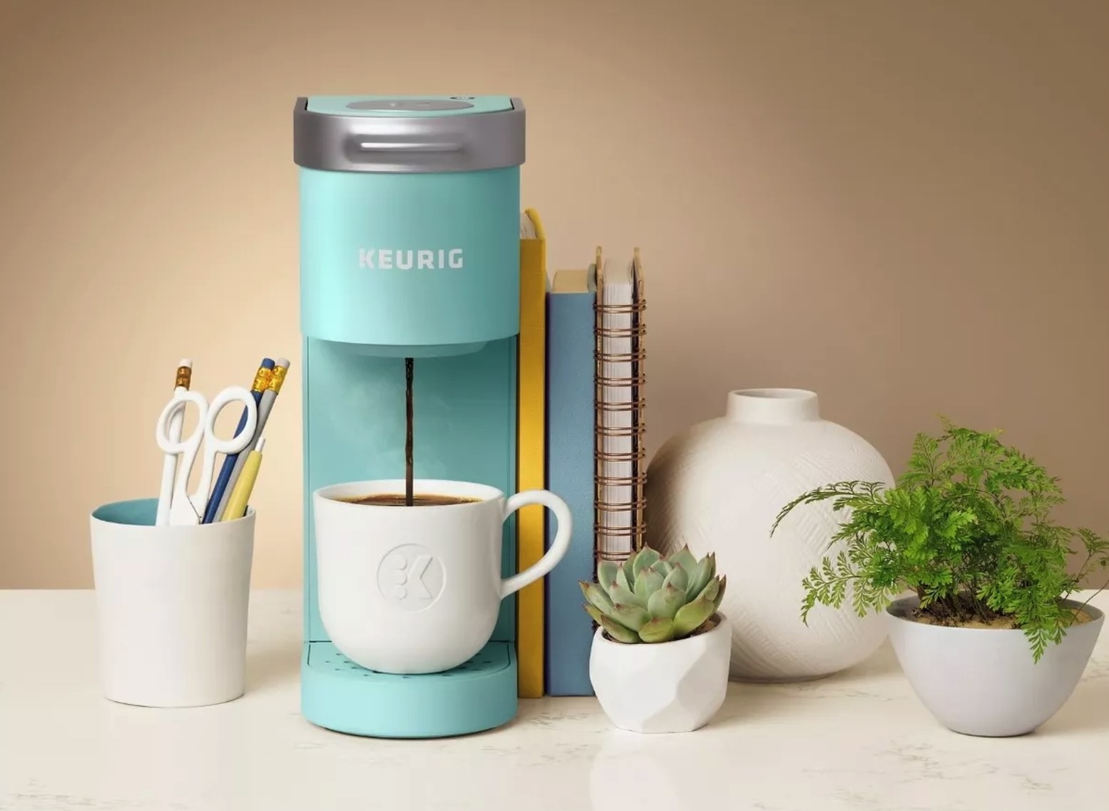 A turquoise Keurig making a cup of coffee next to books, plants, a vase, and a pencil cup.