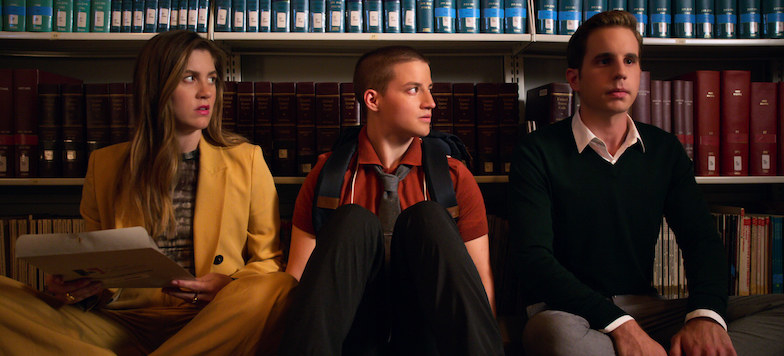 A image from The Politician showing Macafee, James, and Payton sitting on the floor against some bookshelves. Macafee wears a yellow blazer and James wears a red shirt, they both looking at payton in a black sweater looking nervous