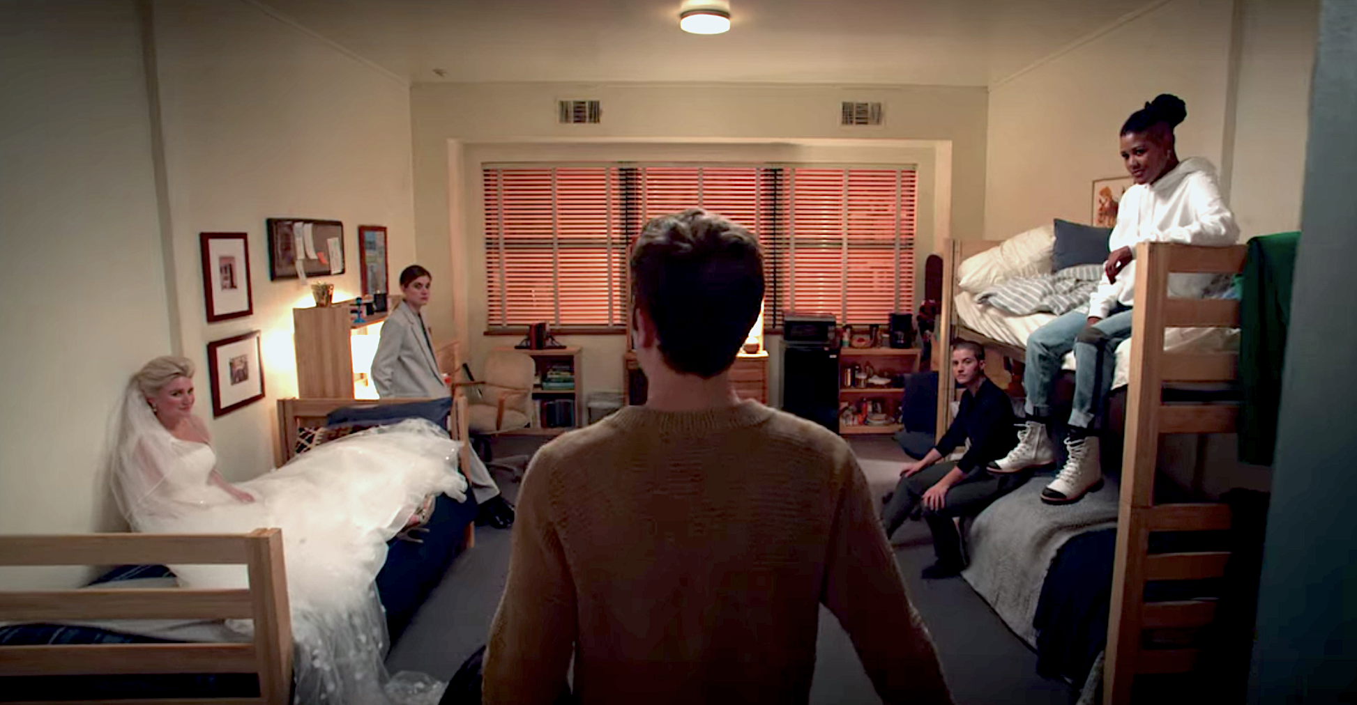 A still from The Politician showing Payton from behind walking into his dorm room and several characters sitting and waiting for him including Macafee, James, Sky, and Alice in a wedding dress