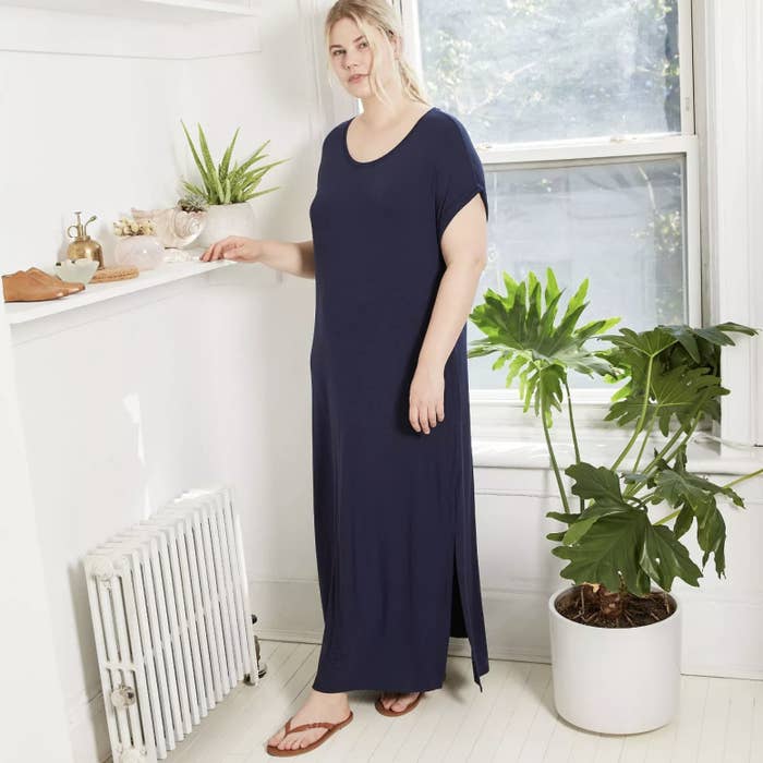 Model is wearing a dark blue short sleeve maxi dress with small slits on the side of the hemline and flip flops
