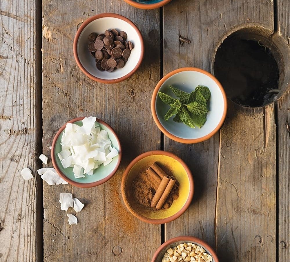 Five small bowls placed on a wooden background Each is filled with a different cooking ingredient like cinnamon, chocolate, mint, and granola