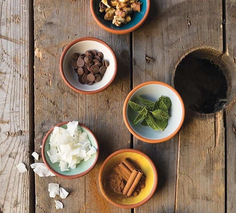 Five small bowls placed on a wooden background Each is filled with a different cooking ingredient like cinnamon, chocolate, mint, and granola