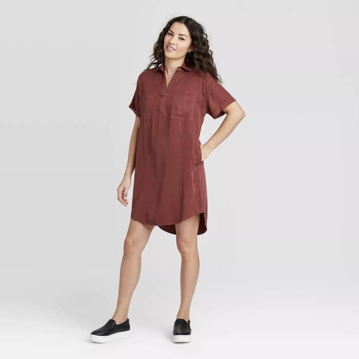 Model is wearing a burgundy short sleeve shirt dress with a collared neckline and black slip-on shoes