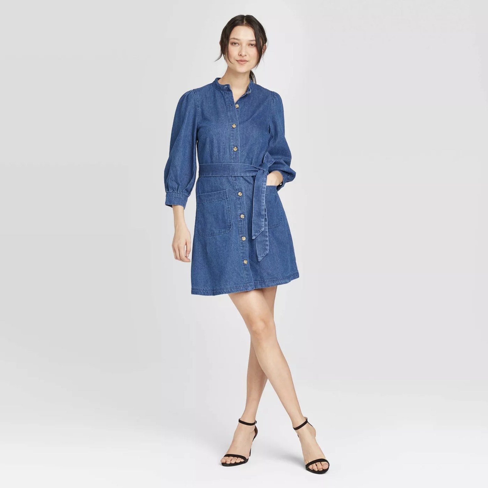 Model is wearing a short three quarter sleeve blue denim dress with buttons down the front and strappy heels
