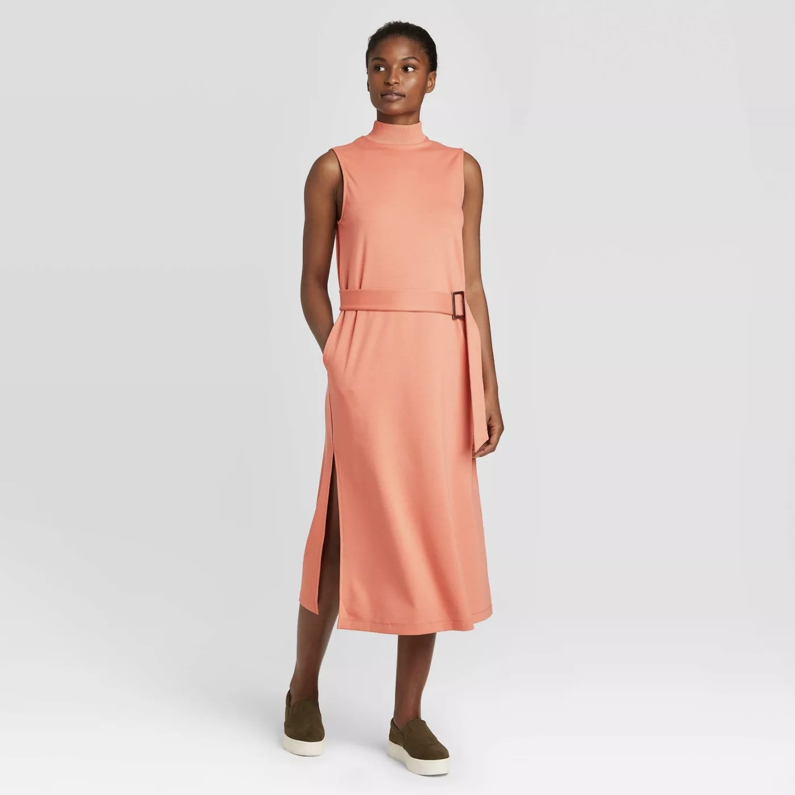 Model is wearing a coral sleeveless midi dress with a turtleneck and slits on the sides of the bottom with black slip-on shoes
