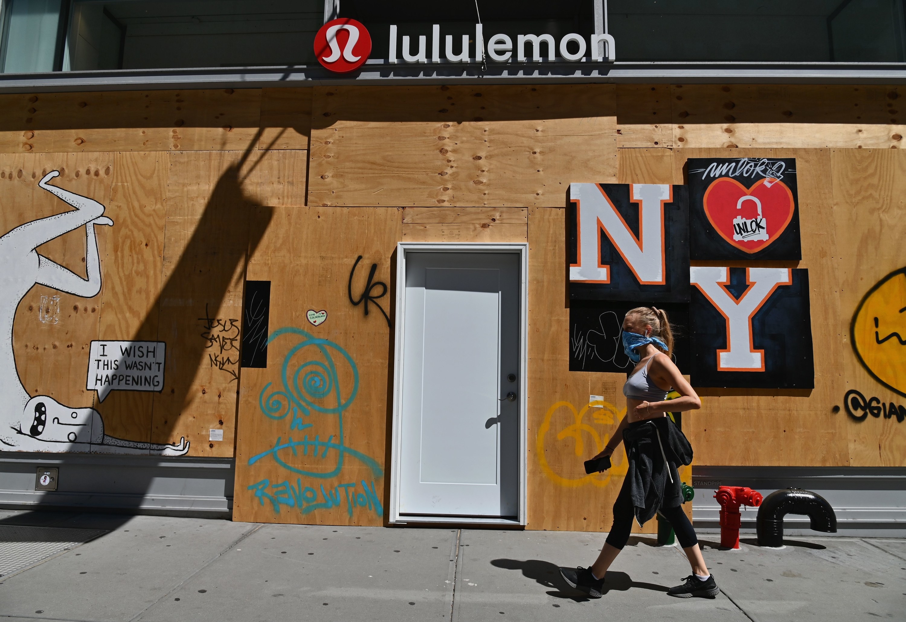 A woman wearing a face-covering walks past a boarded-up Lululemon storefront with a poster attached to it featuring a heart and &quot;NY&quot;