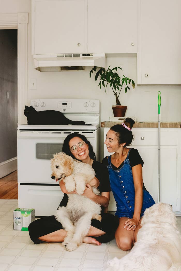 Two women sitting in a kitchen. One is holding a medium-sized white dog in her lap, another one is laying beside one of the girls and a cat is sitting on top of the stove top.