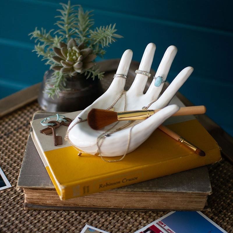 White ceramic hand with rings on the fingers, necklaces draped through the palm and a makeup brush sitting on it