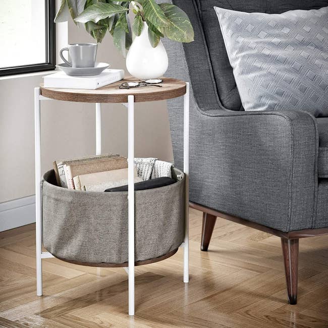 The circular side table with wood-like top, white legs, and a grey fabric bin with books and a blanket in it