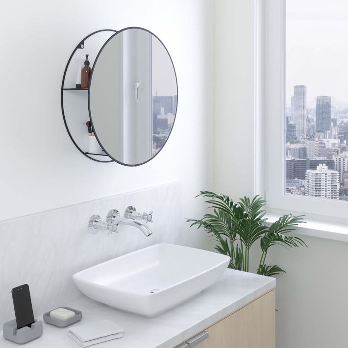 A circular mirror with shelves behind it with assorted bathroom products in it