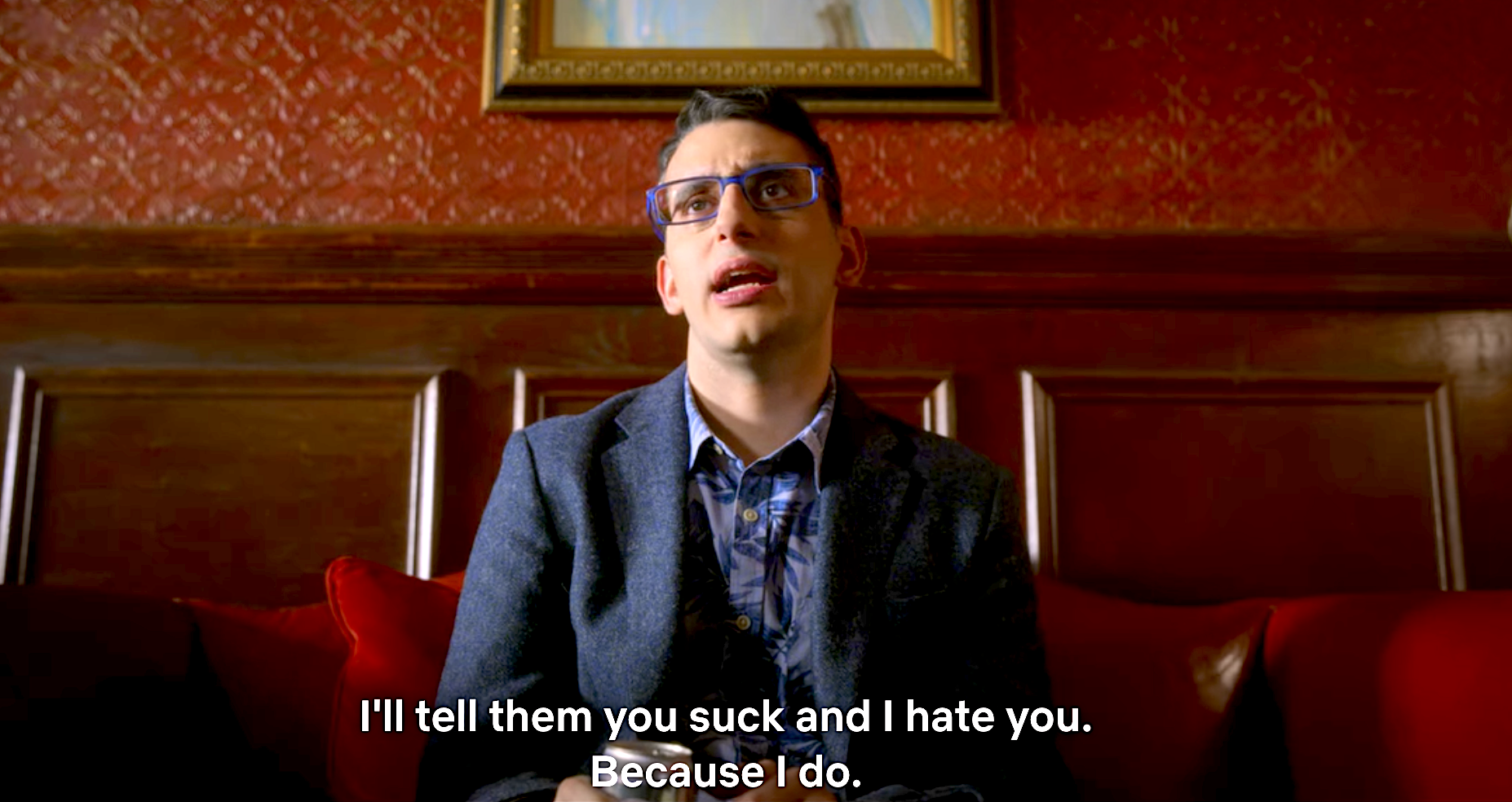 A still from The Politician showing Andrew sitting down in Payton&#x27;s office against a red and mahogany wall in a blue suit saying I&#x27;ll tell them you suck and I hate you because I do