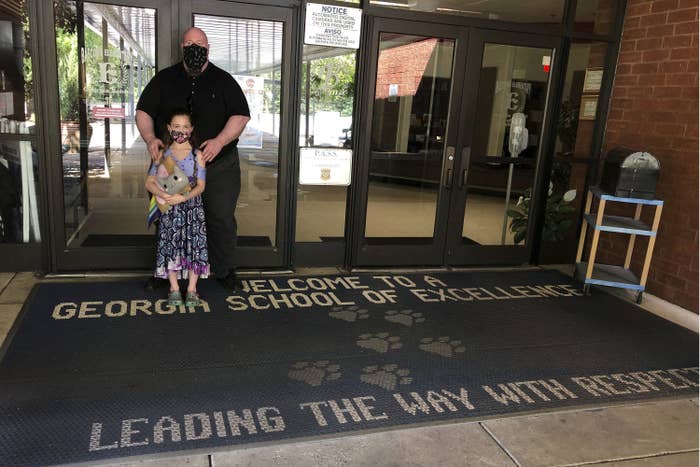 A father wearing a mask stands behind his daughter who is also wearing a mask, on a rug that says &quot;Welcome to a Georgia School of Excellence. Leading the way with respect&quot;