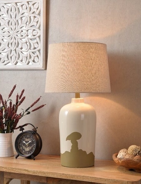 A porcelain table lamp with an ivory glaze on top and a textured olive worn and weathered pattern on the bottom that shifts up to the center of the lamp. The shade is a classic shape in ivory.