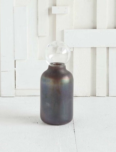 An iridescent gray decorative object in the shape of an old milk bottle with a glass orb on top of the tapered opening 