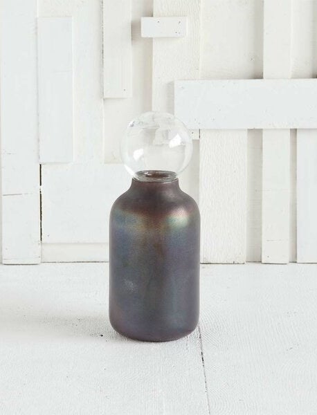 An iridescent gray decorative object in the shape of an old milk bottle with a glass orb on top of the tapered opening 