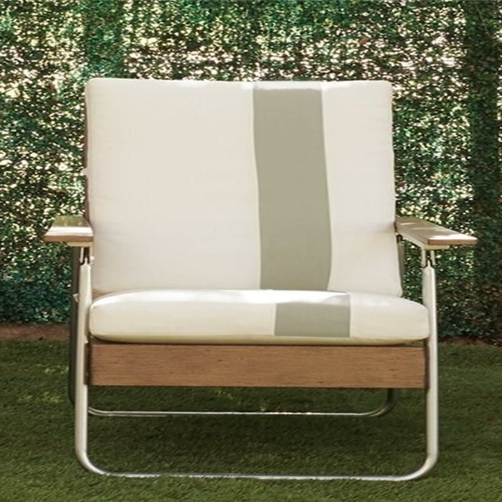 A padded outdoor chair with a wooden detail at the bottom of the seat and matching wooden arm rests. The legs of the chair are gold piping and rounded at the bottom. 