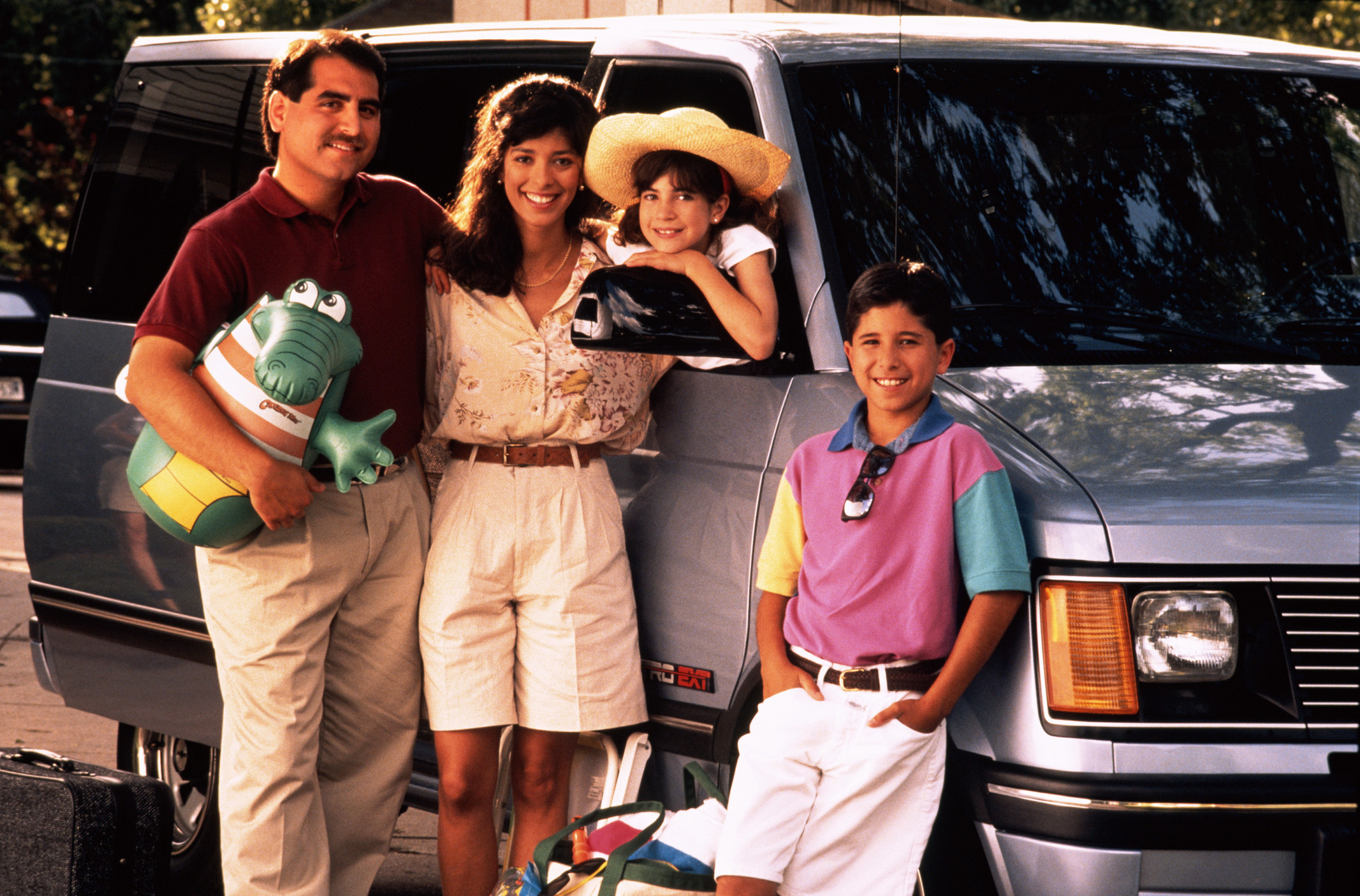 A stock photo of an early &#x27;90s Latinx family posing in front of a grey mini-van as they are about to go on vacation. 