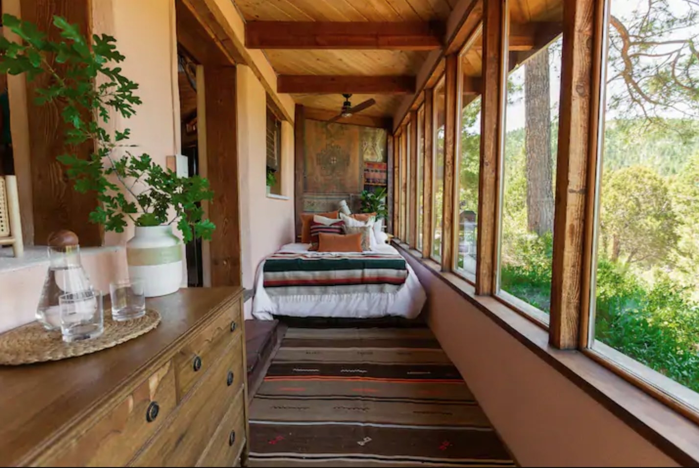 A bed dressed in warm-toned pillows sits nestled in a narrow space between a wall and a large glass window that extends all the way across the room and looks out into the forest.