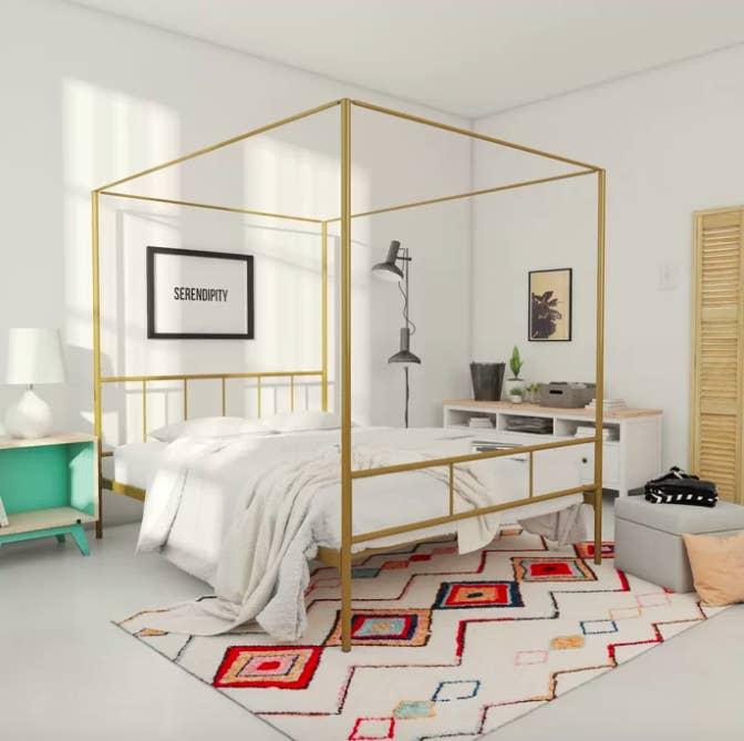 Gold-colored canopy bed with white sheets on top of a rainbow geometric woven rug