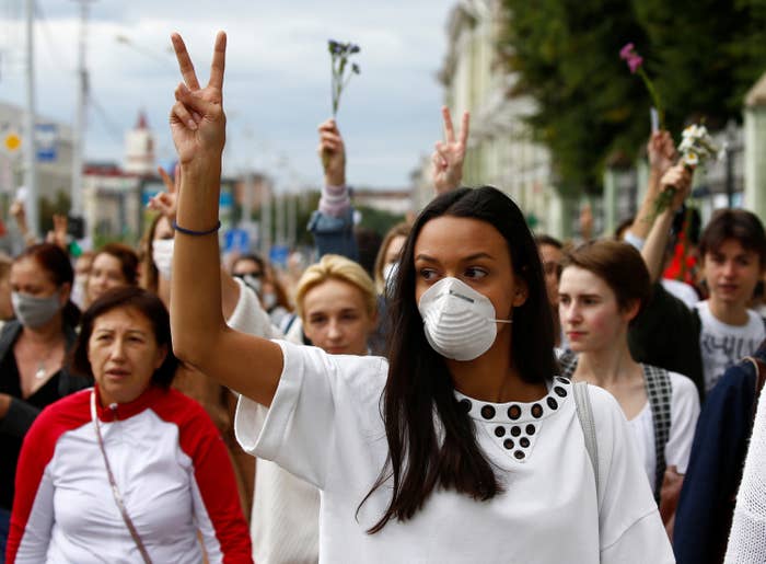 A group of women, many wearing white, march down a street and hold up peace signs with their hands