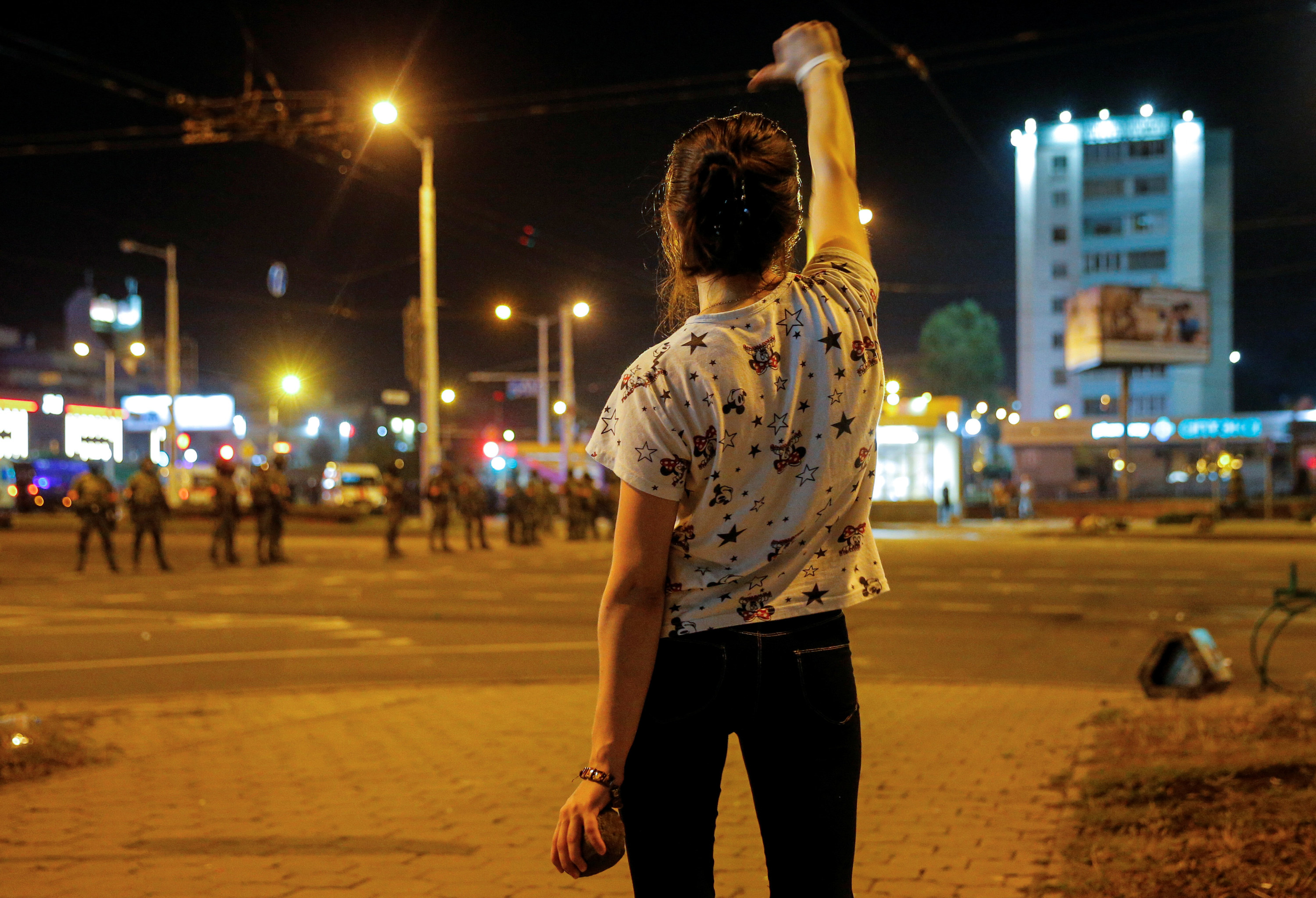 A woman stands in the foreground with her back to the camera and her arm in the air giving a thumbs-down toward a line of law enforcement officers in the background