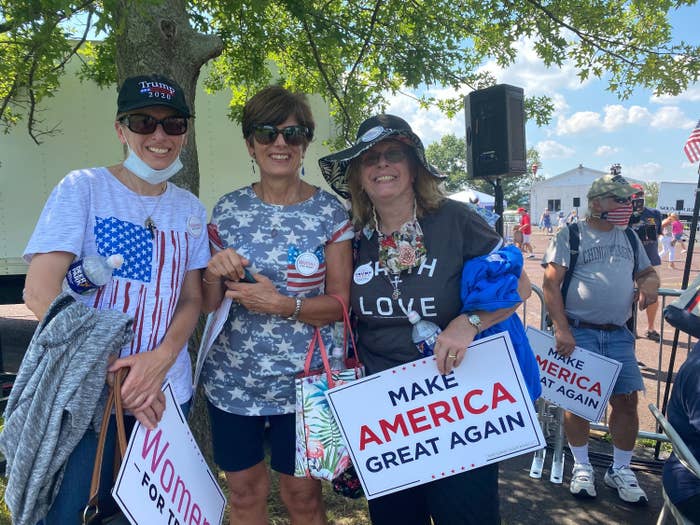 Three white women, two without face masks and one with hers on her chin, hold &quot;Make America Great Again&quot; and &quot;Women for Trump&quot; signs. 