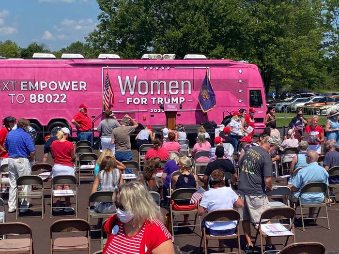 A huge bus painted with &quot;Women for Trump 2020&quot; and other slogans is parked in front of rows of folding chairs occupied by men and women, many of whom are wearing face masks.