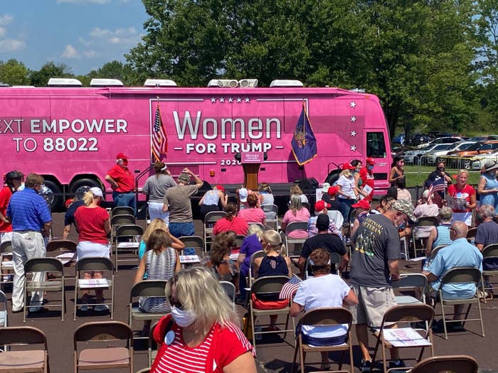 A huge bus painted with "Women for Trump 2020" and other slogans is parked in front of rows of folding chairs occupied by men and women, many of whom are wearing face masks.