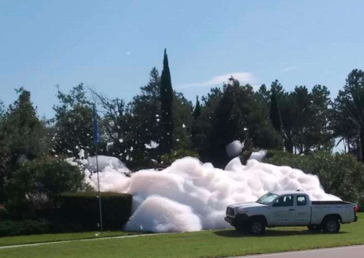 A large flood of foamy suds pours onto grass from a fountain