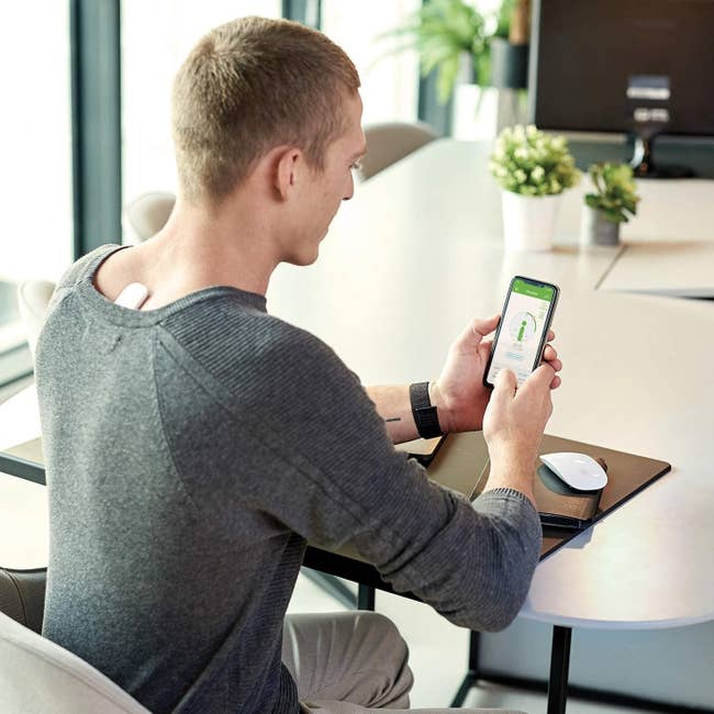 A model wearing finger-length, rectangular posture corrector in the middle of their upper back, between their shoulder blades, as they sit at a desk and check the app on their phone
